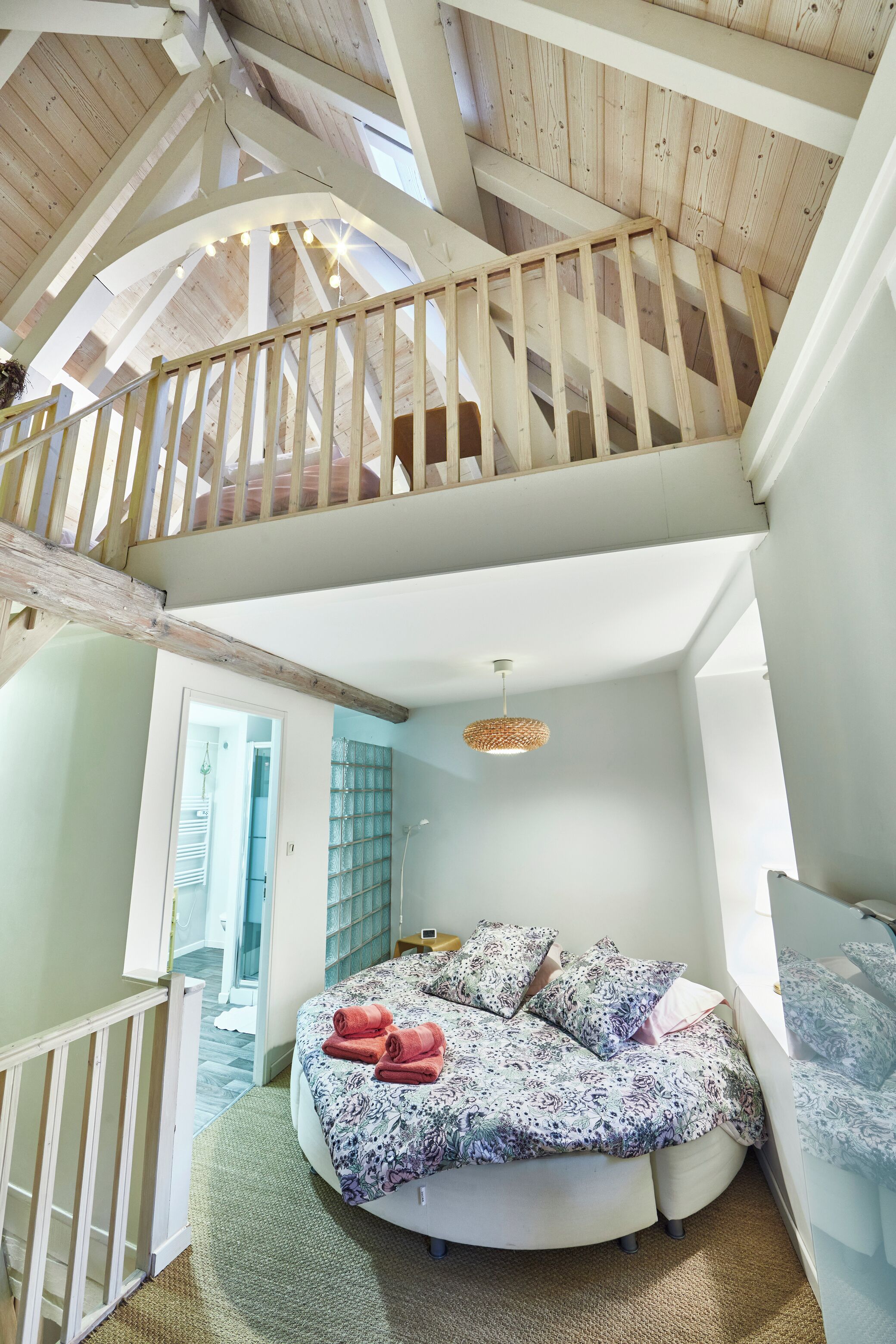 A bedroom with a circular bed, a wooden staircase, and a view of a room with a hanging light fixture.