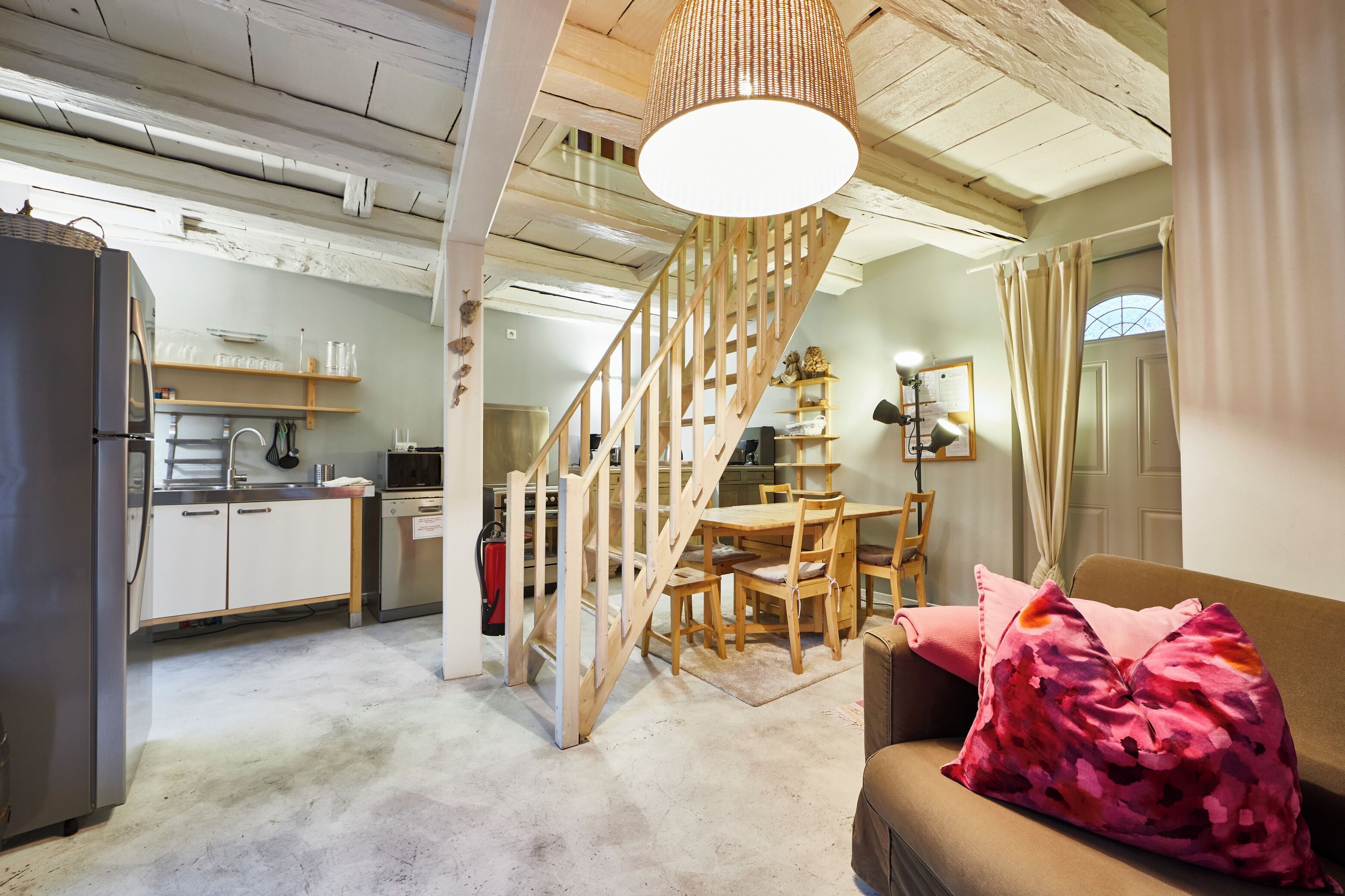 A kitchen with white cabinets, a wooden staircase, a dining area with a table and chairs, and a living room with a sofa and a pink pillow.