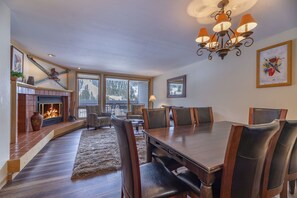 A dining area with a wooden table and chairs, a fireplace, and large windows.