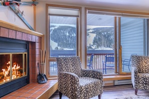A cozy room with a fireplace, two patterned armchairs, and large windows offering a view of a snowy landscape.