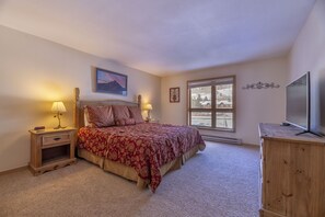 A bedroom with a large bed, two bedside tables with lamps, a TV on a wooden cabinet, and a window with a view of a snowy landscape. - Big 2Br/2 Bath Condo (1443 Sq. Ft.; 2 Living Rooms; Sleeps 10); Walk To Lifts. (Keystone)