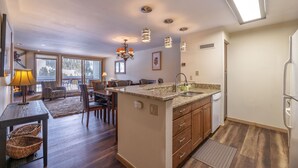 A modern kitchen with a granite countertop, wooden cabinets, and a built-in island. There is a dining area with a table and chairs, and a large window providing natural light. - Big 2Br/2 Bath Condo (1443 Sq. Ft.; 2 Living Rooms; Sleeps 10); Walk To Lifts. (Keystone)