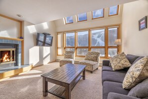 A cozy living room with a fireplace, comfortable seating, large windows with a mountain view, and a flat-screen TV mounted on the wall.