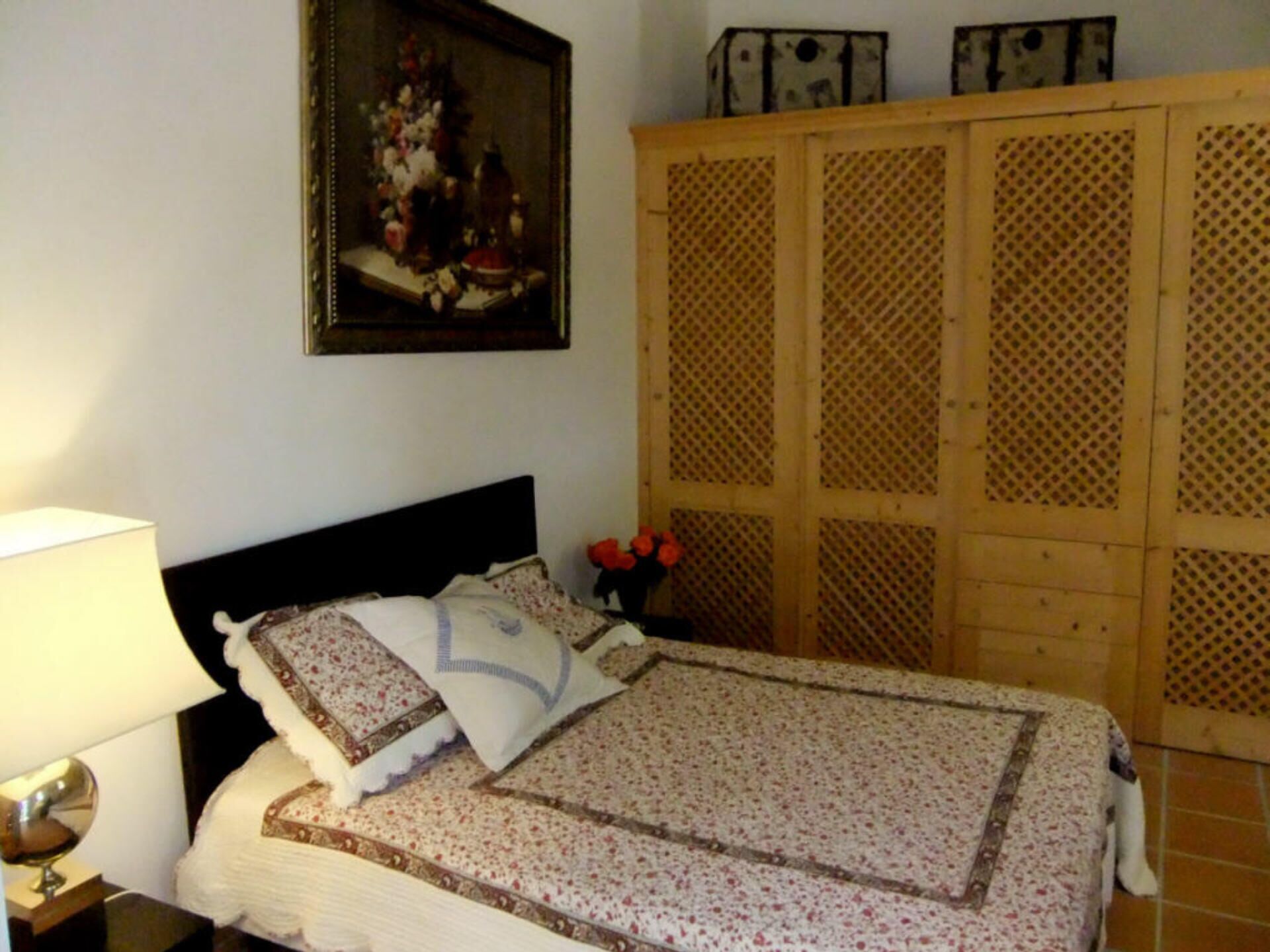 A bedroom with a bed, a nightstand, a lamp, a wooden wardrobe, and a painting on the wall.