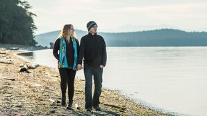 Zwei Personen gehen an einem Strand entlang, mit einem See und Bergen im Hintergrund.