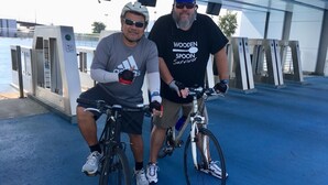 Dos personas en bicicleta en un peaje con un suelo azul y un edificio moderno al fondo.