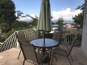 Ein Balkon mit rundem Tisch und Stühlen, einem Sonnenschirm und Blick auf das Meer und die Berge.