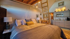 A bedroom with a large bed, wooden ceiling beams, a ladder, and a kitchenette in the background.