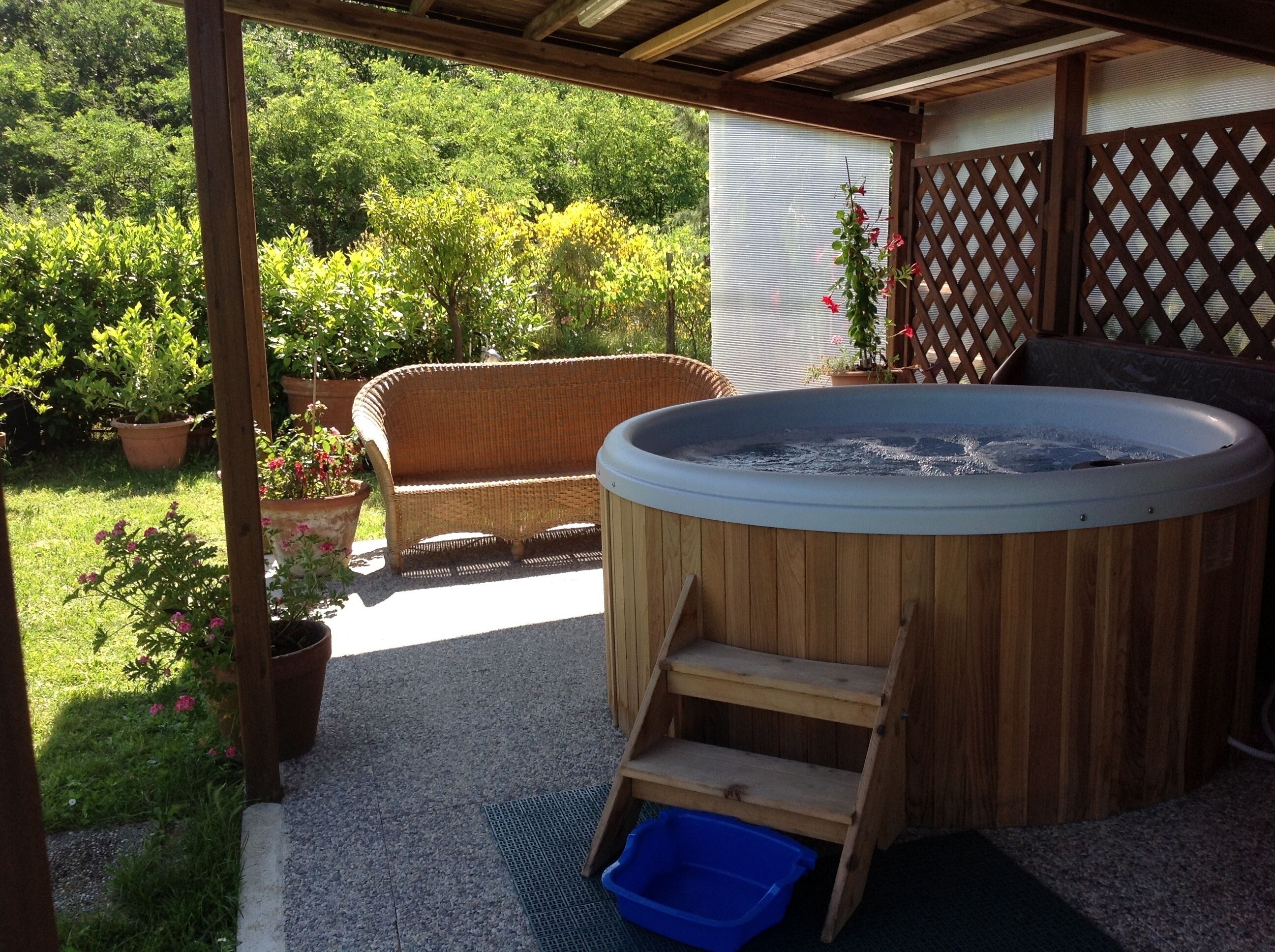 A hot tub with wooden steps, a wicker sofa, and potted plants in a covered outdoor area.