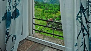 Una habitación con un ventanal grande, cortinas blancas con estampado floral y vistas a un paisaje verde con montañas al fondo.