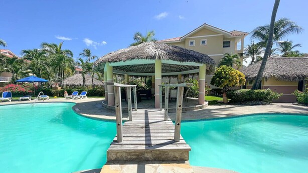A pool area with a wooden deck leading to a covered seating area, surrounded by palm trees and a building in the background. - Charming Studio in Ocean Dream (Cabarete)