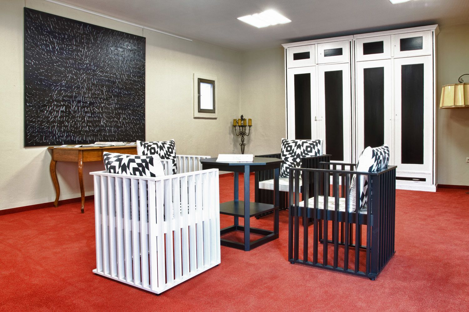 A room with a red carpet, a white crib, a black and white patterned chair, a wooden table, and a large black abstract painting on the wall.