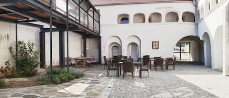 Un patio con suelo de piedra, mobiliario exterior y un edificio con arcos y tejado de tejas rojas.