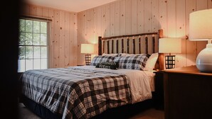 A bedroom with a wooden paneled wall, a large window, a bed with plaid bedding, two bedside lamps, and a nightstand with a lamp.