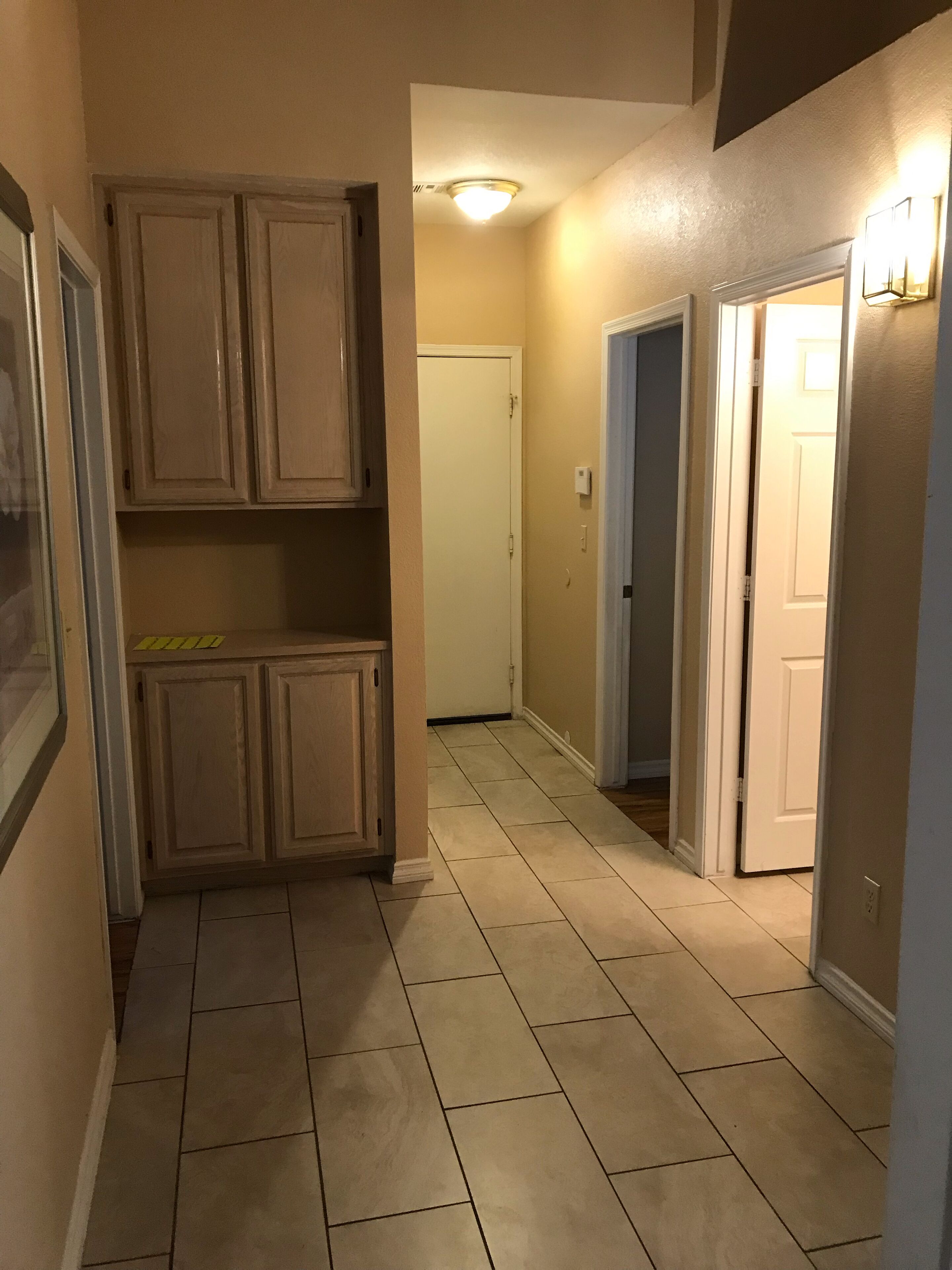 A narrow hallway with beige walls and a tiled floor. There are wooden cabinets on the left and a door at the end.
