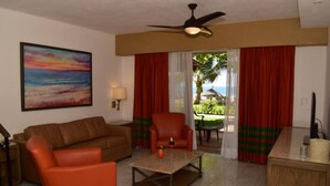 A living room with a brown sofa, an orange armchair, a coffee table, and a ceiling fan. There is a painting on the wall and a view of the outdoors through a window with red curtains. - Deluxe Beach Front Villa “Tesoro D” (Ixtapa-Zihuatanejo,)