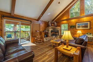 Un intérieur de chalet chaleureux avec une cheminée en pierre, des meubles en bois et un balcon offrant une vue sur la forêt.