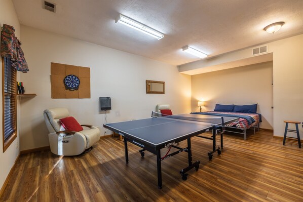 Une pièce équipée d’une table de ping-pong, d’un fauteuil de massage, d’un jeu de fléchettes et d’un lit recouvert d’une couverture bleue et rouge.