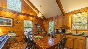 A wooden cabin interior with a dining table, chairs, a bench, a kitchen area, and a large window. - Bluebird Cabin (Hocking Hills area) (Sugar Grove)