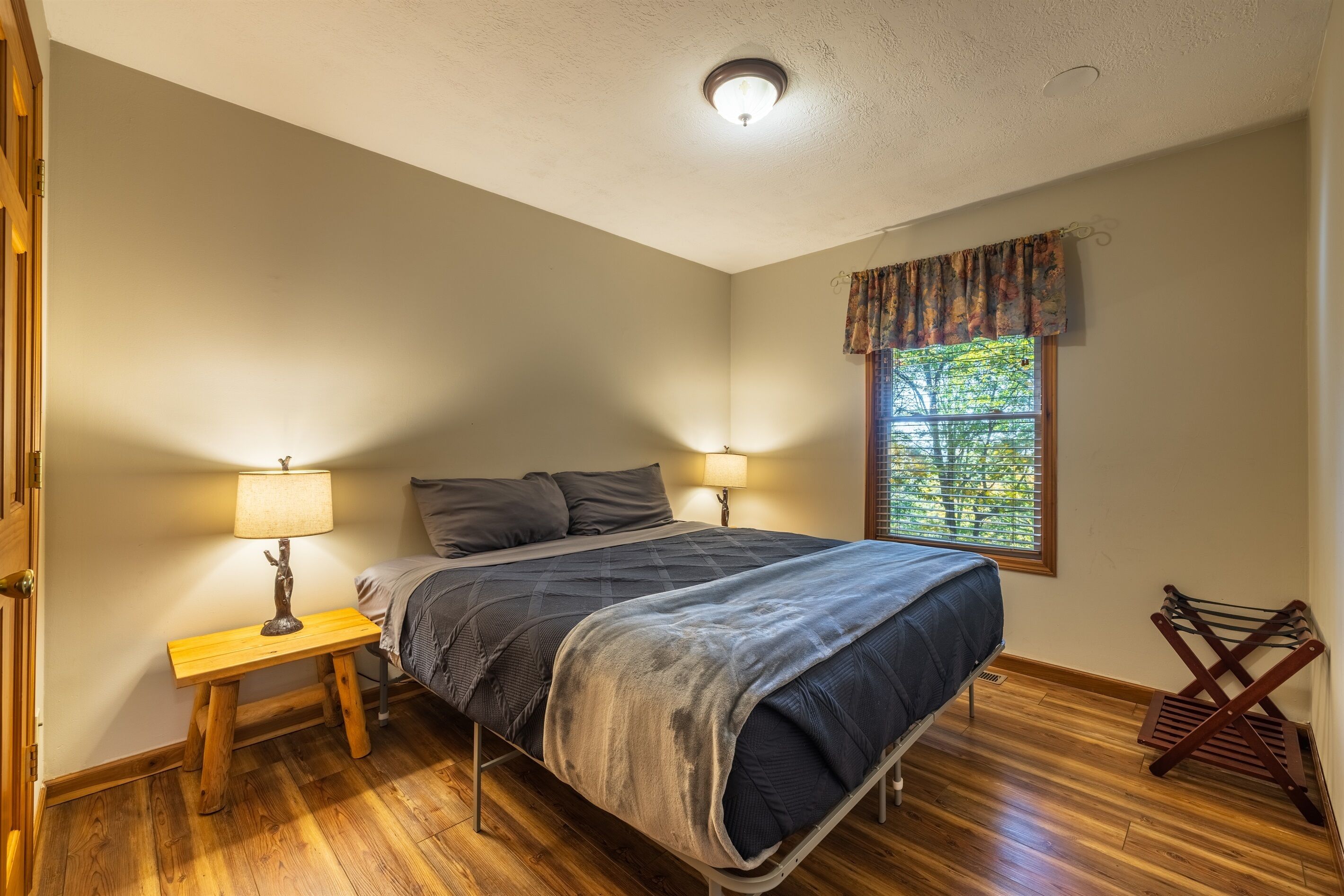 A bedroom with a bed, two bedside lamps, a window with a view, and a wooden floor.