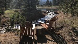 A rustic outdoor seating area with two wooden chairs and a table, overlooking a river and surrounded by trees. - Look No Further! Brand New Build in Winter Park/Fraser (Fraser)