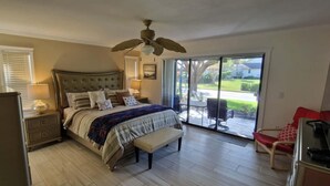 Ein Schlafzimmer mit Bett, Nachttischen, einer Bank, einem Deckenventilator, einer Schiebetür zu einer Terrasse und einem roten Sessel.