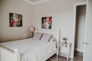 Une chambre avec un lit blanc, deux tableaux accrochés au mur, une table de chevet avec une lampe et une porte donnant sur une autre pièce.