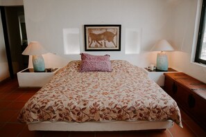 Une chambre avec un grand lit, deux lampes de chevet, un tableau encadré au mur et un banc en bois près de la fenêtre.