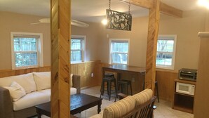 A living room with a sofa, a wooden coffee table, a striped armchair, a kitchen area with a microwave and toaster, and a hanging decorative piece.