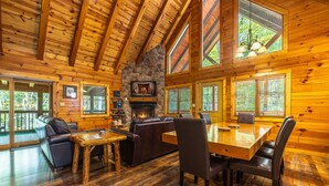 A log cabin interior with a stone fireplace, wooden dining table, leather sofas, and large windows. - Cardinal Cabin (Hocking Hills area) (Sugar Grove)