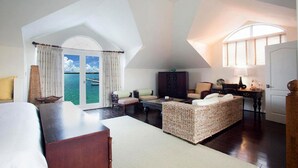 A spacious room with a large window offering a view of the sea, a cozy seating area with a woven ottoman, and a wooden desk with a lamp. - 3 Bedroom 3.5 Bath Villa on the Water (Exuma)