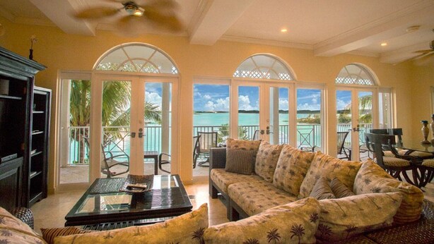 A spacious living area with a sofa, a coffee table, and a TV. There are two doors leading to a balcony with a view of the ocean. - 3 Bedroom 3.5 Bath Villa on the Water (Exuma)