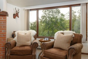 A room with two brown leather armchairs with white fur throw pillows, a wooden side table with a picture frame, and a large window with a view of trees.