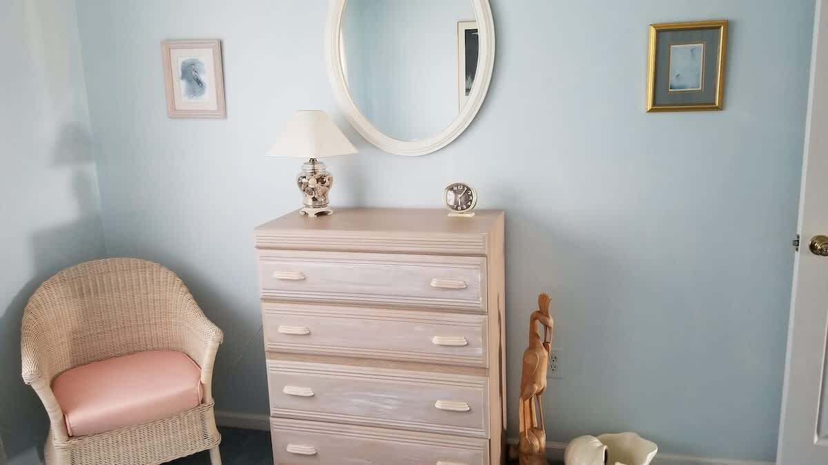 A room with a wicker chair, a dresser with drawers, a round mirror, and two framed pictures on the wall.