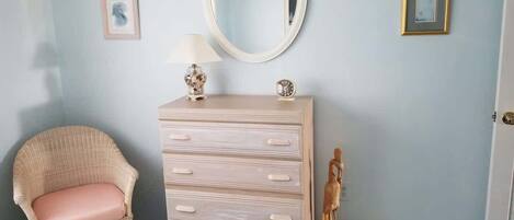 A room with a wicker chair, a dresser with drawers, a round mirror, and two framed pictures on the wall.