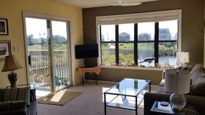 Un salón con sofá, una mesa de centro, un televisor sobre un mueble bajo, una ventana con vistas a zonas verdes y un balcón.