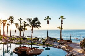 Zona de piscina en un resort, con palmeras, sillas de descanso y sombrillas, con vista al océano.