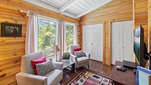 A cozy cabin interior with wooden walls, two armchairs with cushions, a television, and a large window with curtains.