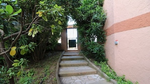 Un escalier en pierre menant à une porte, entouré d’une végétation luxuriante et d’un bâtiment dont le mur présente une texture particulière.