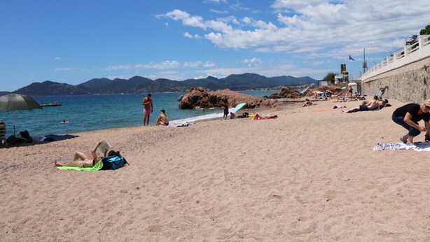 Une plage où des gens se prélassent au soleil et se baignent, un rivage rocheux et des montagnes en arrière-plan.