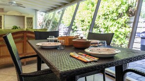 A dining area with wicker furniture, a table set with plates, glasses, and cutlery, and a view of greenery through large windows. - Banyan Tree Cottage  Tropical Siesta Key Retreat - Walk to Sunset at Shell Beach (Sarasota)