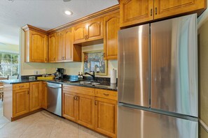 A kitchen with wooden cabinets, a stainless steel refrigerator, and a black countertop. - Banyan Tree Cottage Tropical Siesta Key Retreat - Walk to Sunset at Shell Beach (Sarasota)