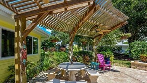 Ein überdachter Patio-Bereich mit Steintischen und -stühlen, umgeben von üppiger Bepflanzung und einer hölzernen Pergola.