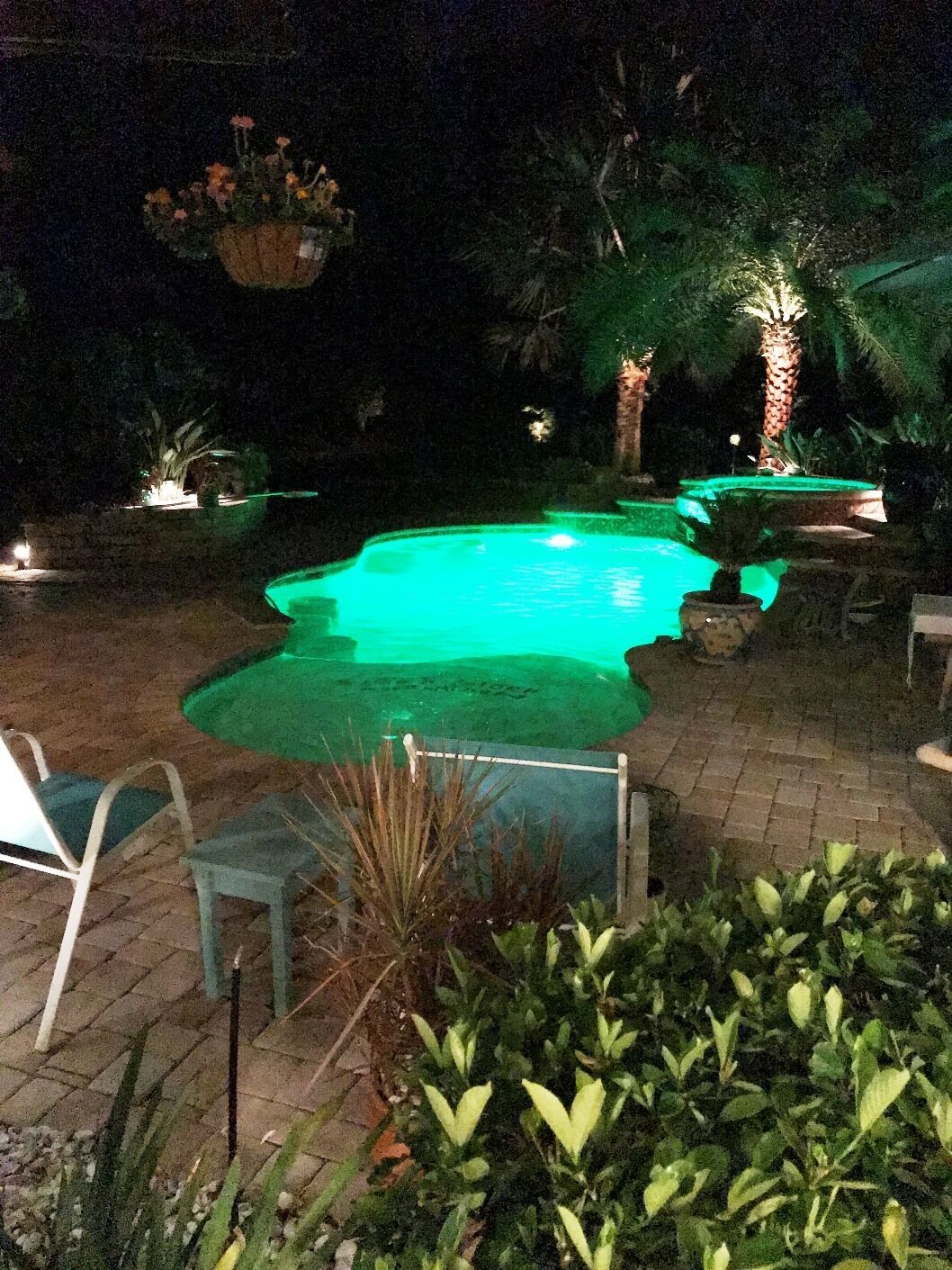 A nighttime view of a swimming pool area with a green-lit pool, surrounding plants, and a few chairs.