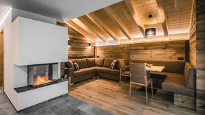 A cozy loft-style room with a built-in fireplace, wooden ceiling beams, and a seating area with a sofa and table. - Lüch de Costa (Corvara in Badia)