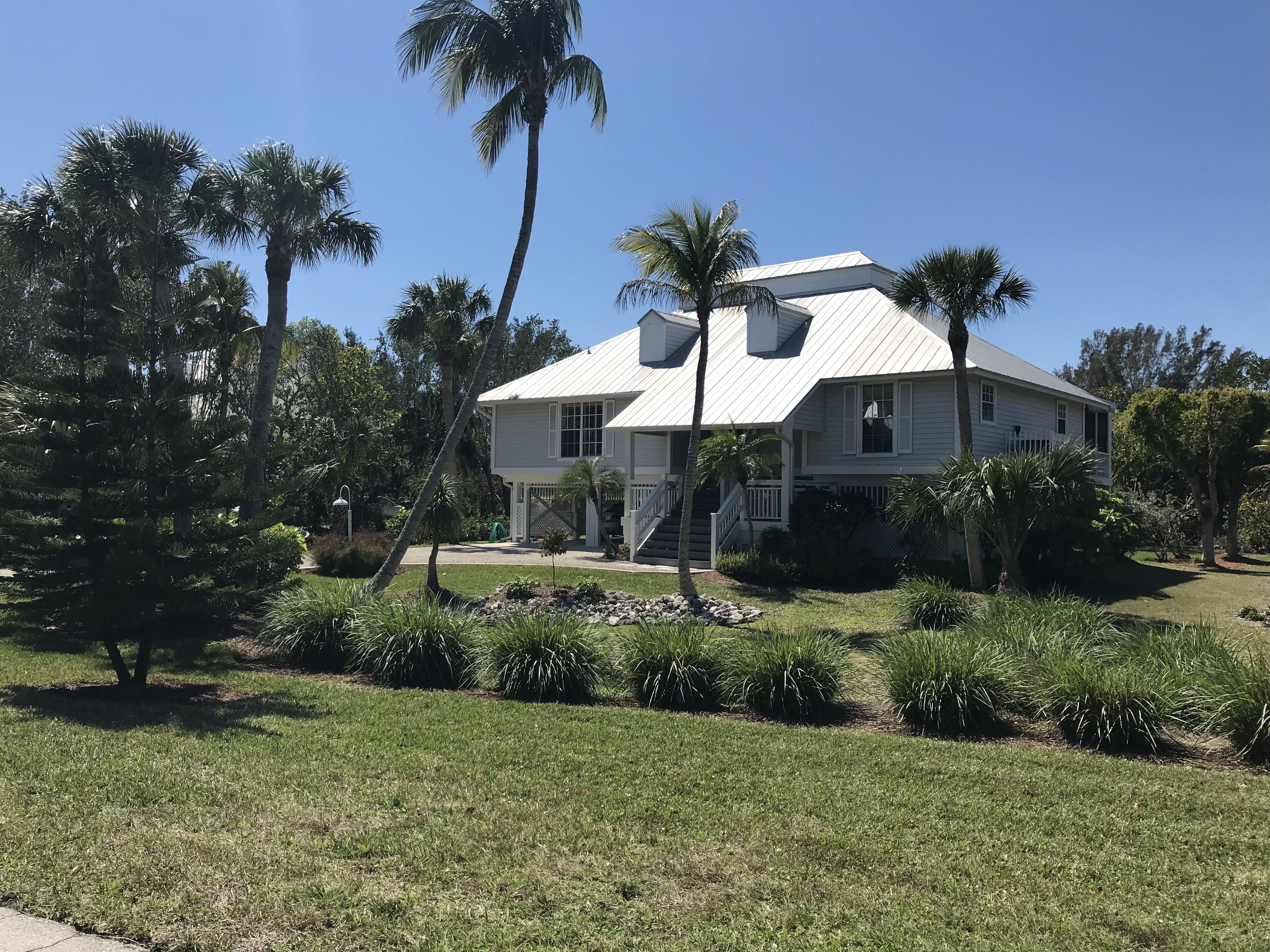 Uma casa de dois andares com fachada branca, varanda coberta e um pequeno jardim frontal com palmeiras e arbustos.