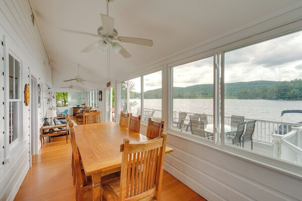 A spacious dining area with a large wooden table and chairs, large windows offering a view of a lake and hills, and ceiling fans.