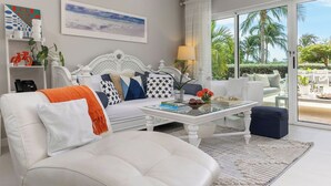 A modern living room with a white sofa, a glass coffee table, and a white armchair. There is a bookshelf with decorative items and a painting on the wall.