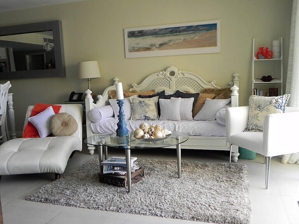 Ein Wohnzimmer mit einem weißen Sofa, einem Glastisch und einem weißen Sessel. An der Wand hängt ein Gemälde und ein Regal zeigt dekorative Objekte.