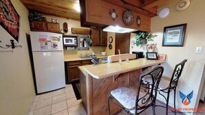 A compact kitchen with wooden cabinets, a white refrigerator, a microwave, and a bar area with two chairs.